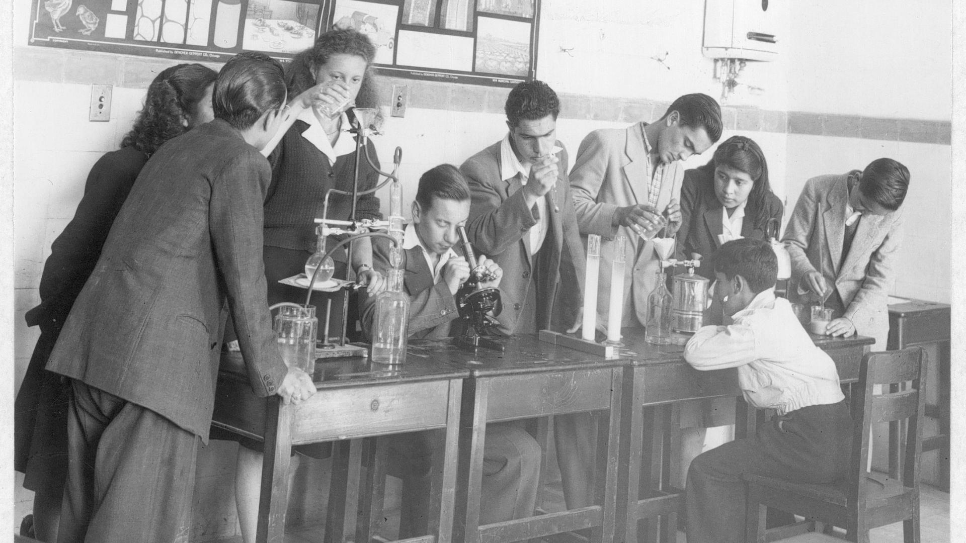 Gurpo de estudiantes escolares, hombres y mujeres, en una sala con material de laboratorio.