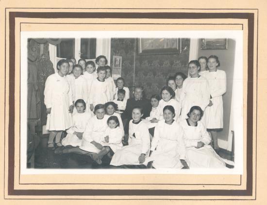 Gabriela Mistral sentada, rodeada de alumnas con delantal blanco.